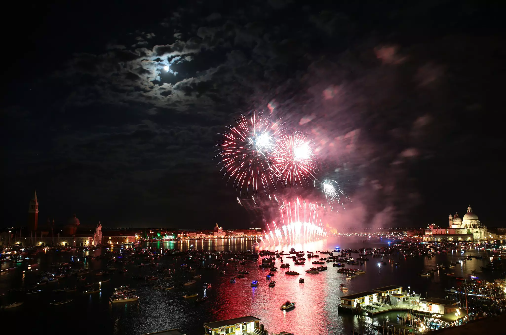 Fireworks explode over a river in a city, with boats moored to watch the display