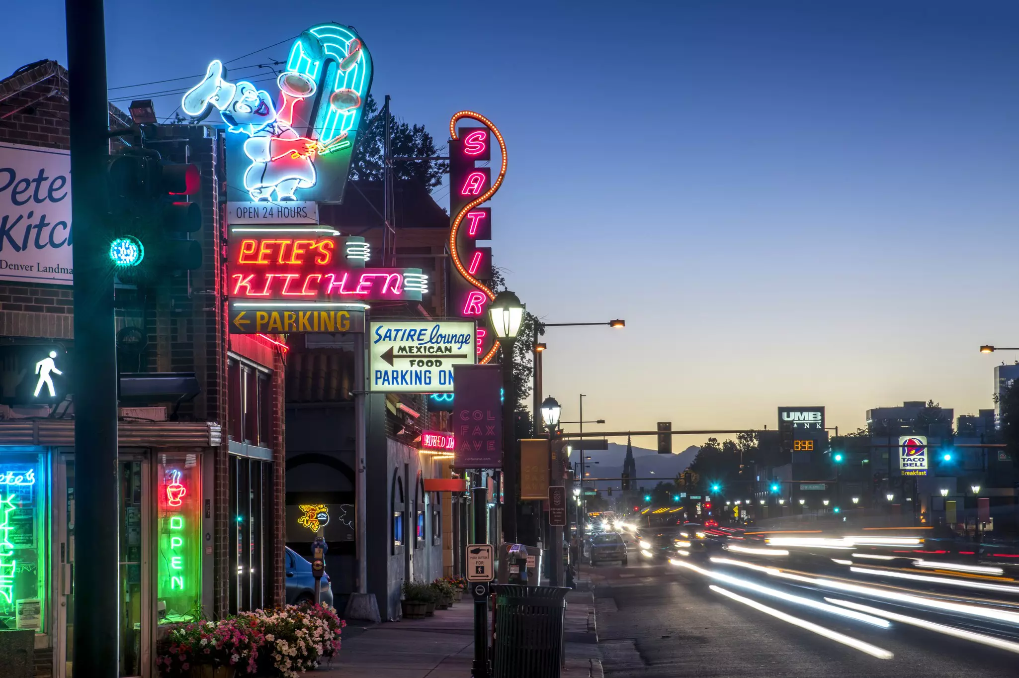 Neon lights from various businesses are illuminated, as blurry car headlights are seen at the right on a city street at dusk.
