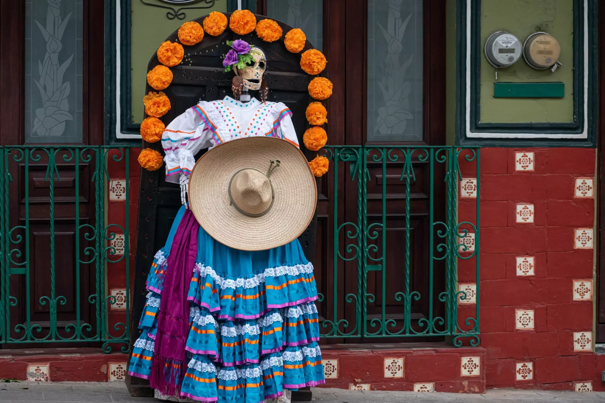 A fake skeleton dressed with traditional clothes, holding a hat and surrounded by large marigolds as decoration for Mexican Day of the Dead.
