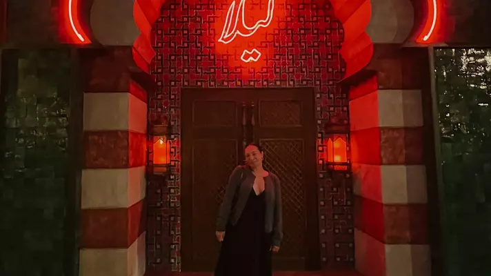 A woman standing outside the lighted entryway to a restaurant at night.