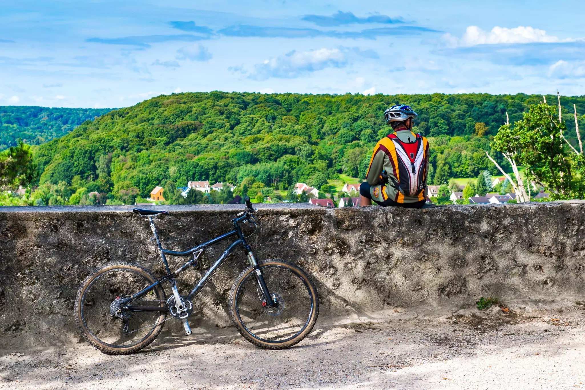 A Cyclist sits on a stone wall facing green scenery; a bicycle is leaning against the wall.