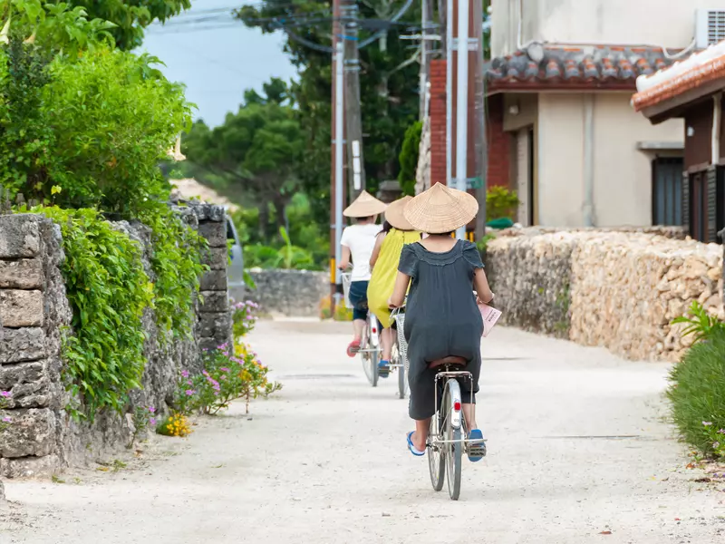 Three people riding bicycles, one after the other, strolling through the traditional village of Taketomi, wearing the traditional straw hat.