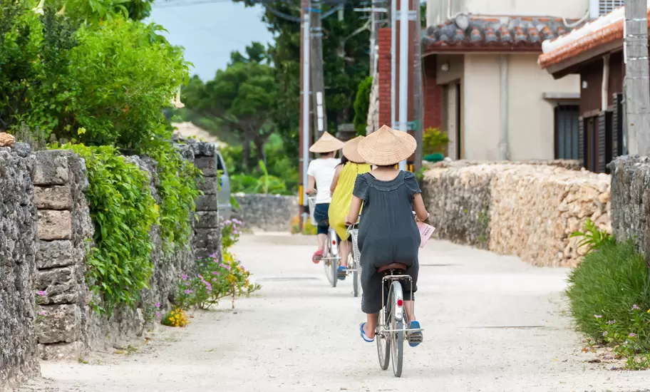 Three people riding bicycles, one after the other, strolling through the traditional village of Taketomi, wearing the traditional straw hat., License Type: media, Download Time: 2026-02-13T17:33:49.000Z, User: nic.dhoedt_lonelyplanet, Editorial: false, purchase_order: 56530 - Guidebooks, job: Global Publishing WIP, client: Lonely Planet Japan 19, other: Nicolas D'Hoedt