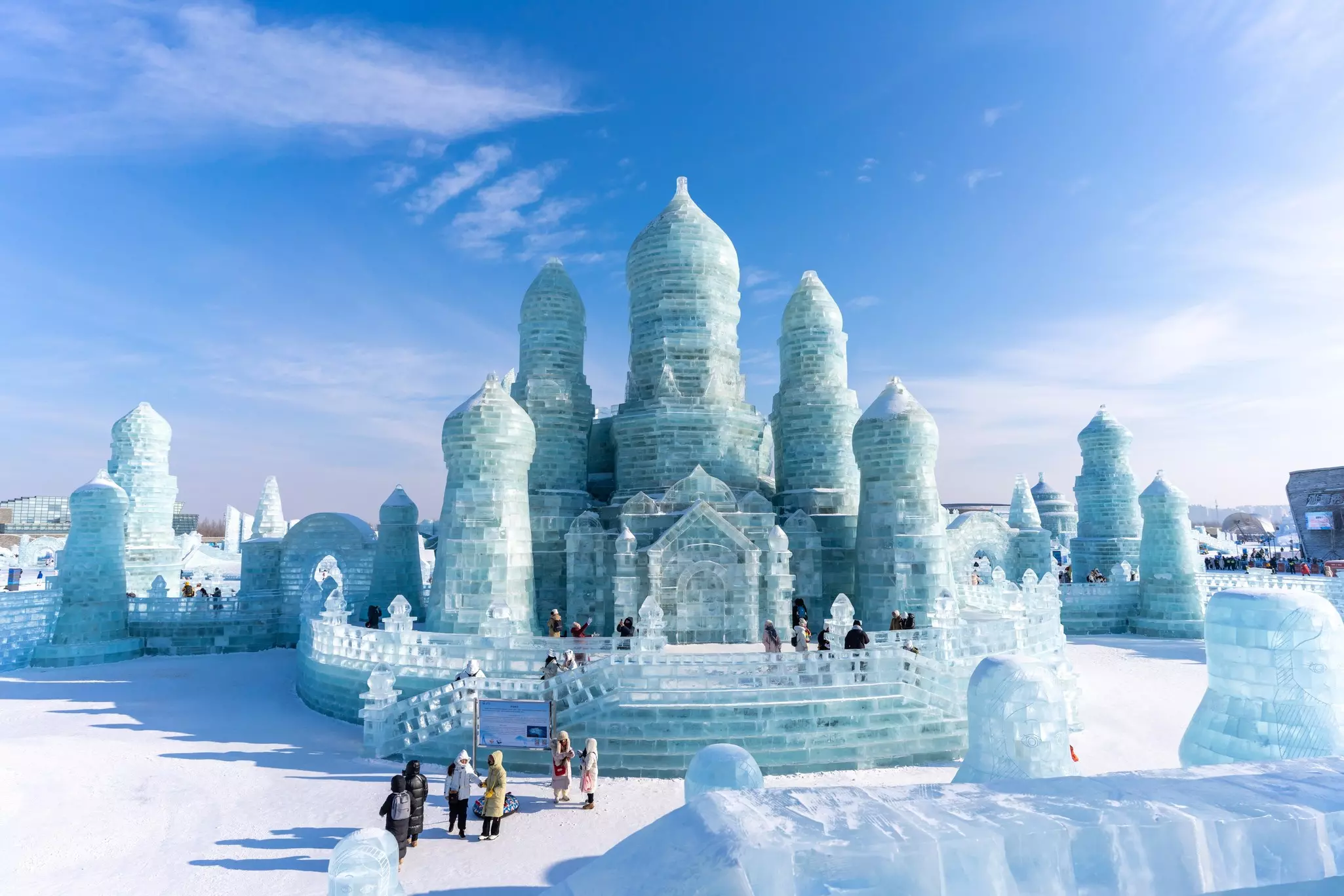 Visitors walk in front of structure shaped like church, complete with towers and domes, carved completely from ice, under a bright sky.