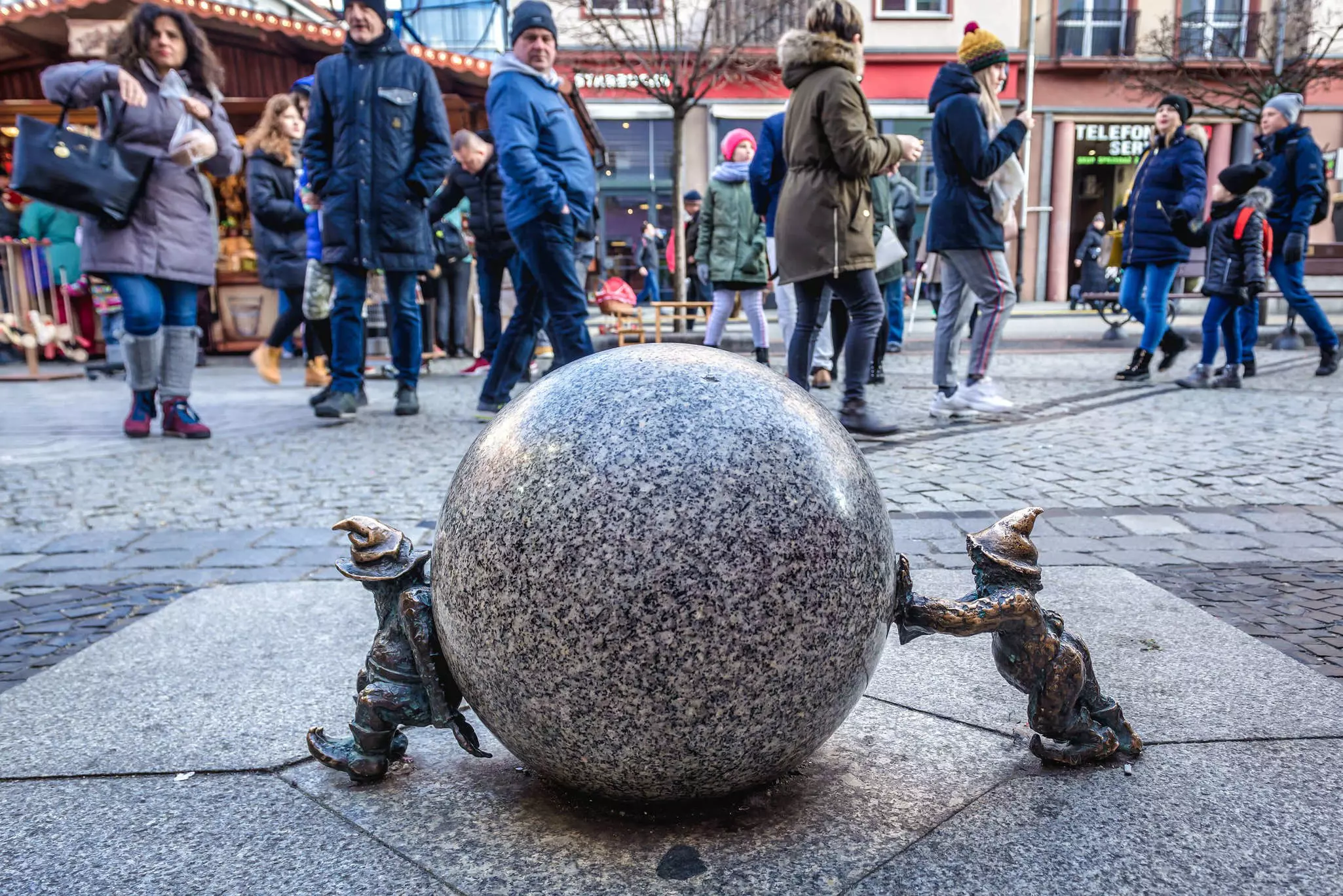 People looking down at a statue of two small figures on either side of a round marble ball, both pushing inward.