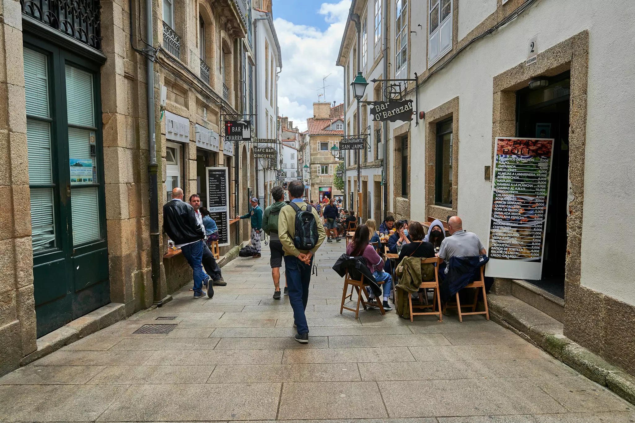 People standing, walking, and sitting in an alleyway between buildings on a sunny day.