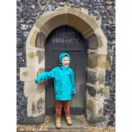 A boy in a blue jacket with the hood up smiles under an arch; above him a sign reads "No Nuisance."