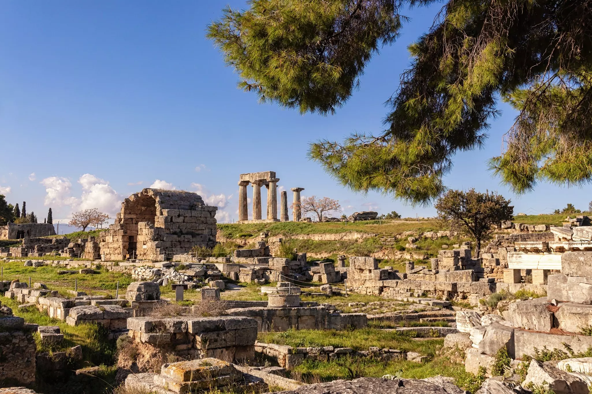Columns are seen in the distance at an archaeological site filled with ruined stone structures and trees.