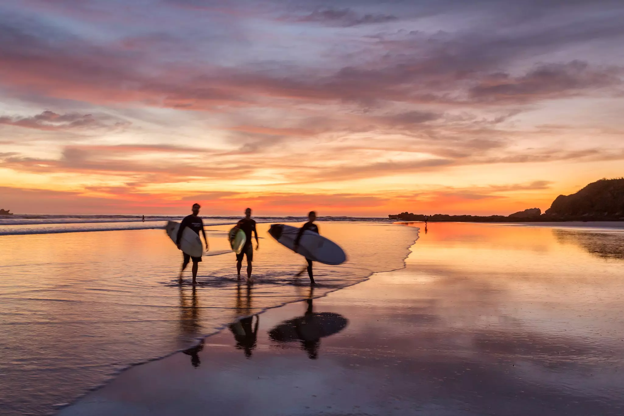 Surfers walk along a beach shore at sunset.