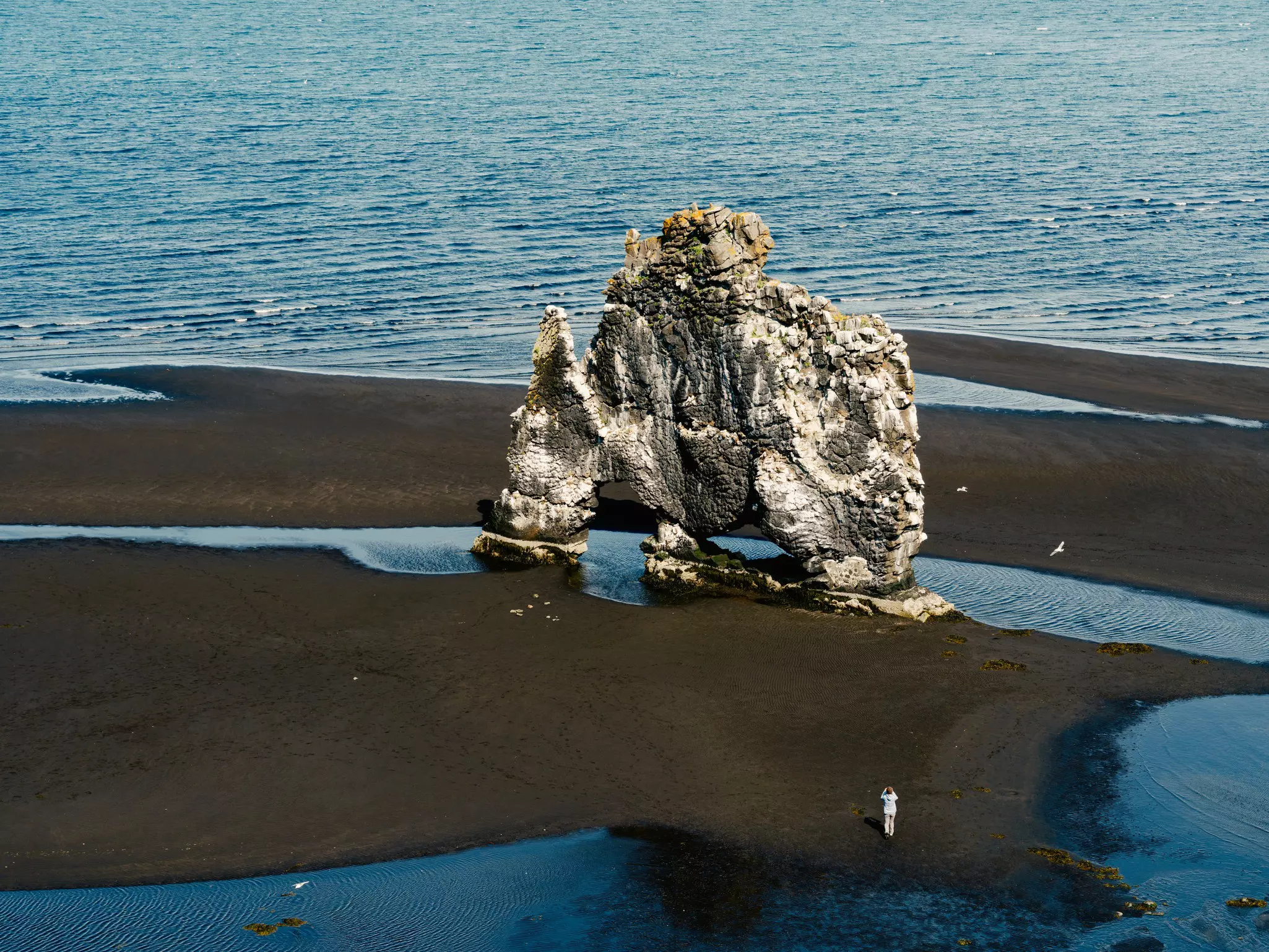 Aerial shot of a person on a dark sandy beach taking a photo of a sea stack with the water in the distance on a partly sunny day.