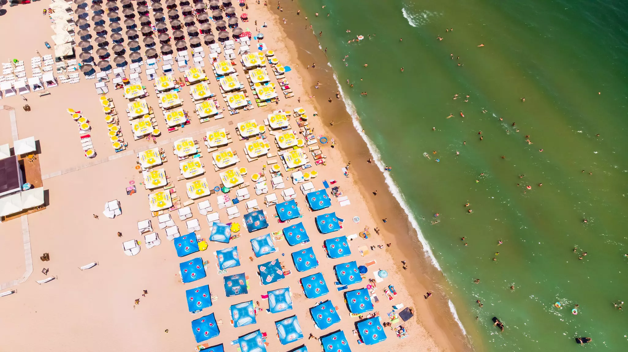 Aerial shot of many square blue and yellow umbrellas and rounded grass umbrellas on sandy beach with greenish sea to the right.