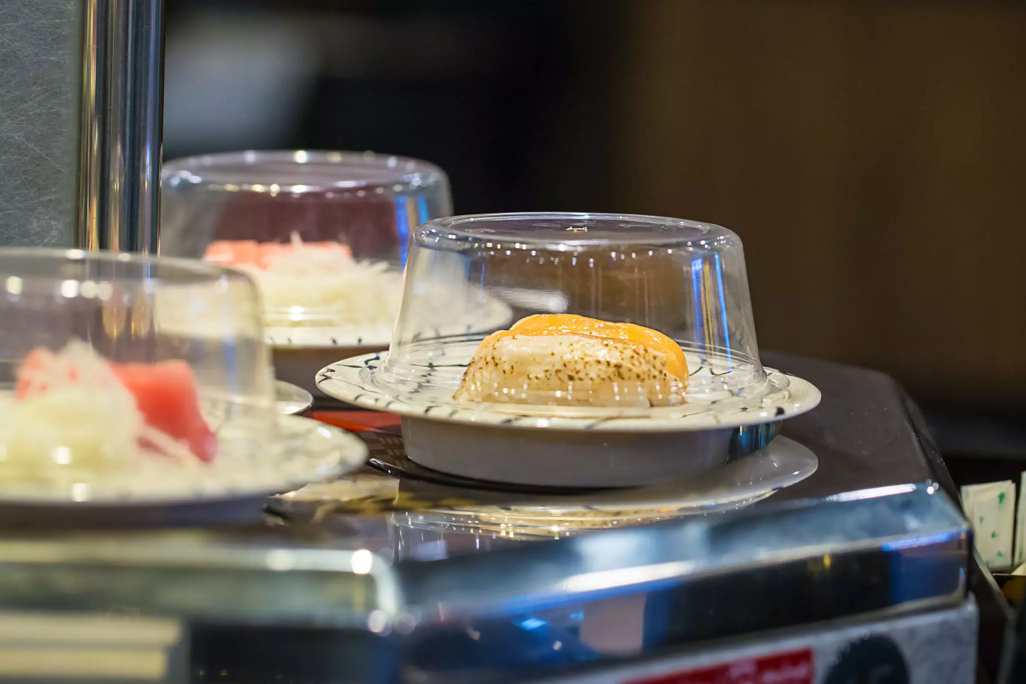 Three covered dishes on plates on a revolving belt in a restaurant