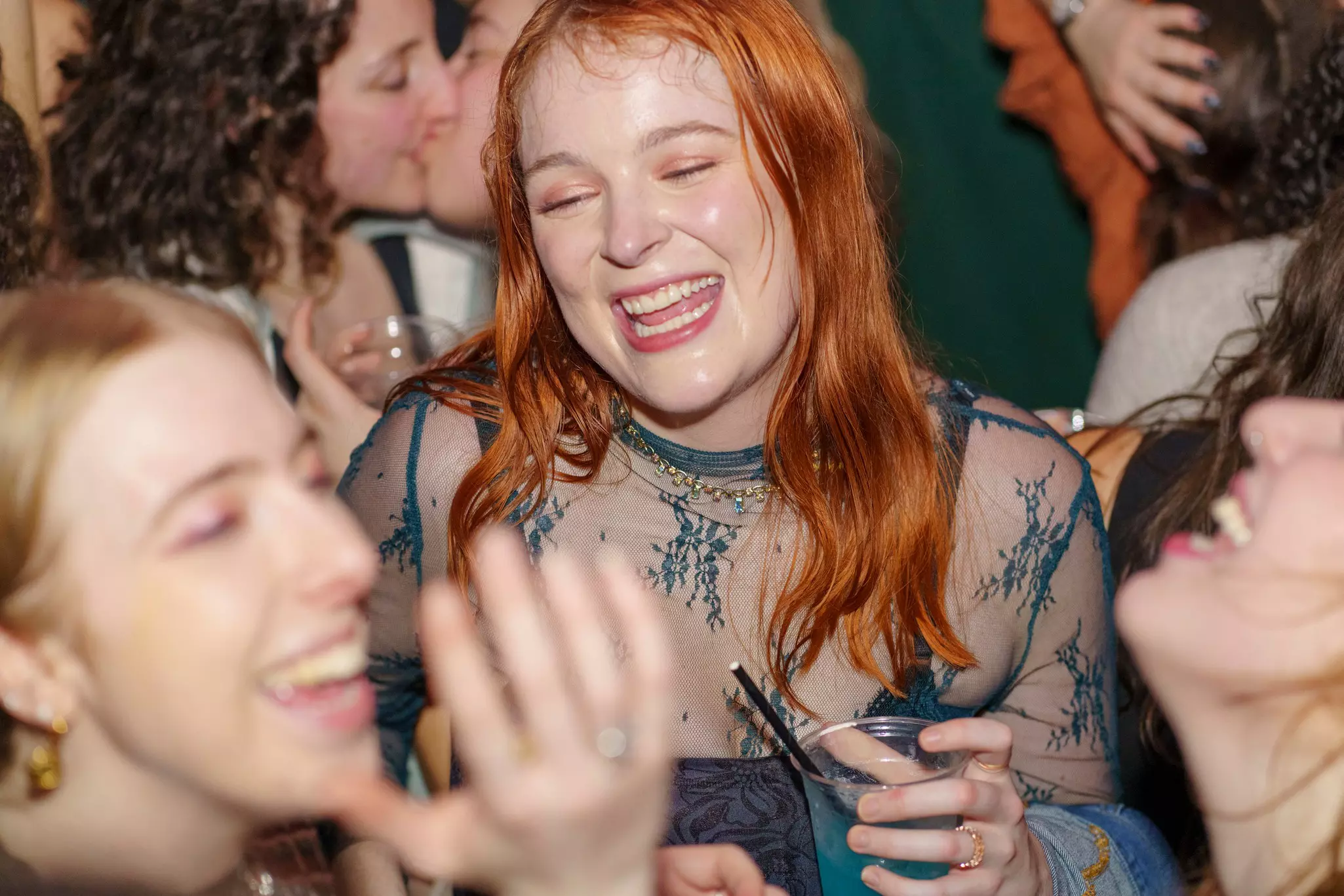 A smiling woman with red hair holds a drink in the center of a crowd.