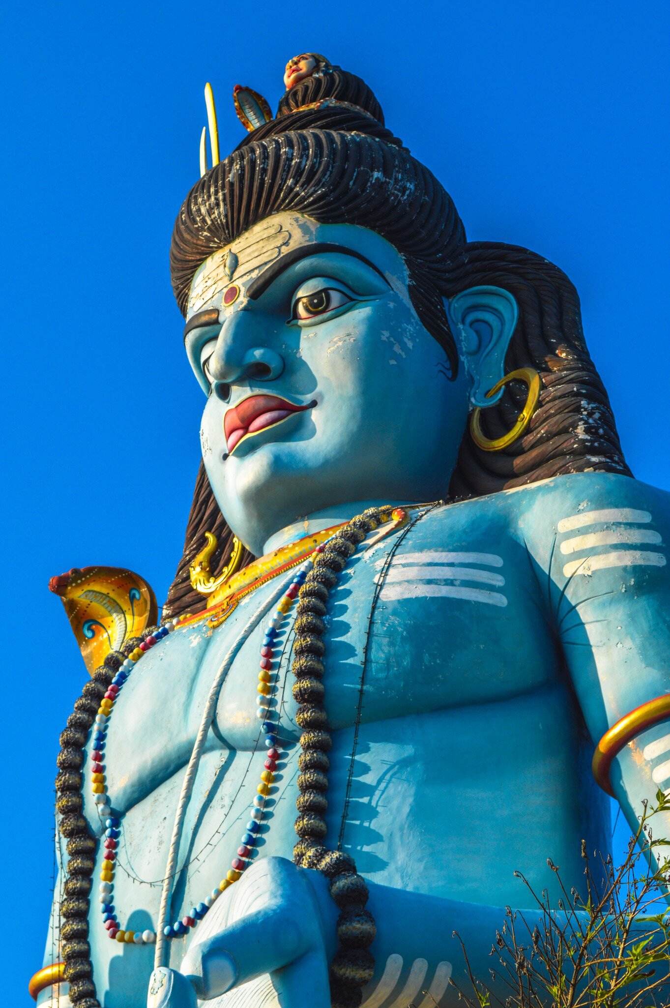 Blue-skinned figure with dark coiled hair, gold hoop earrings and strings of beads, seen from below against deep blue sky