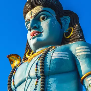 Blue-skinned figure with dark coiled hair, gold hoop earrings and strings of beads, seen from below against deep blue sky