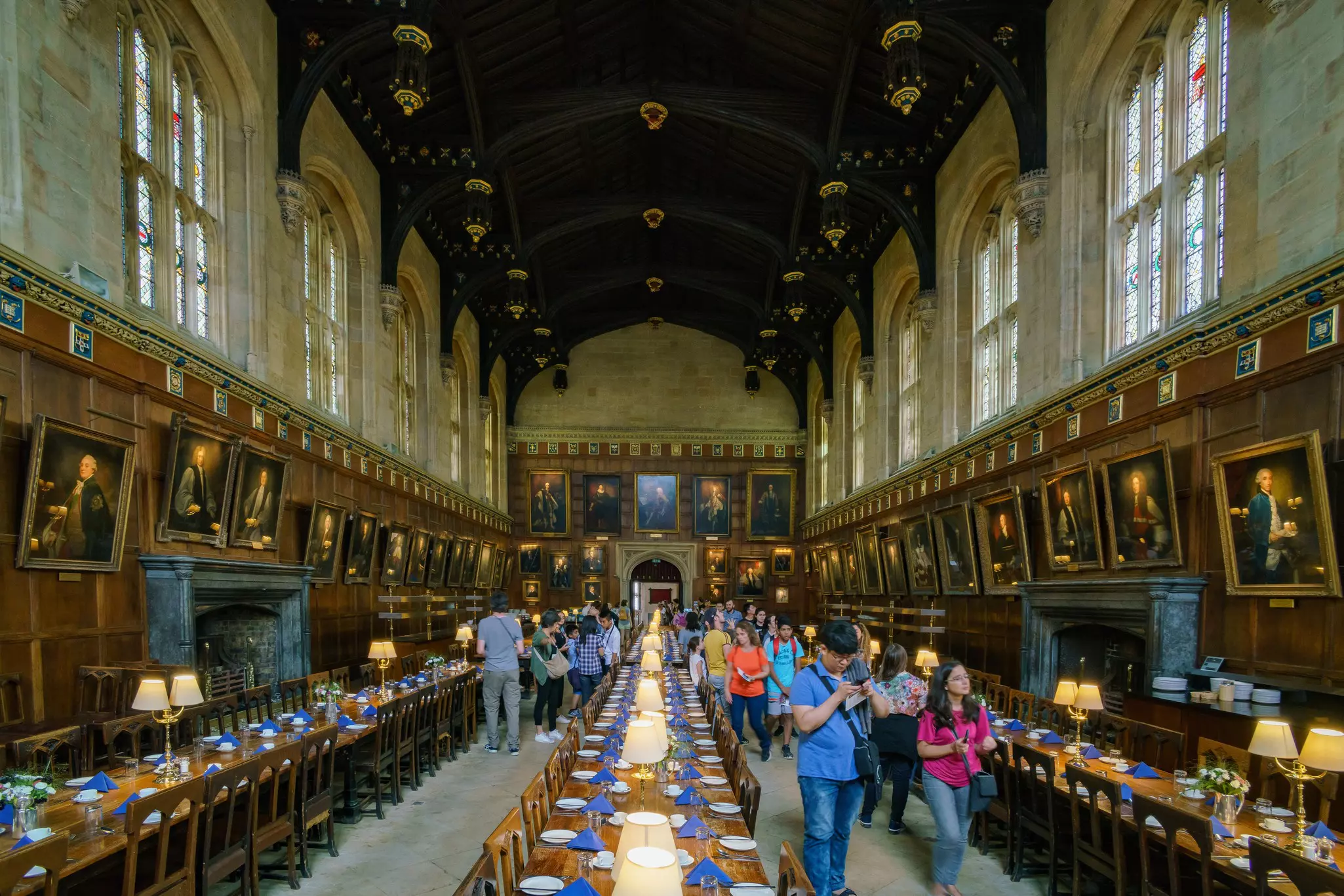 Tourists walk through a large dining hall with ornate ceiling and historic portraits along wooden walls.