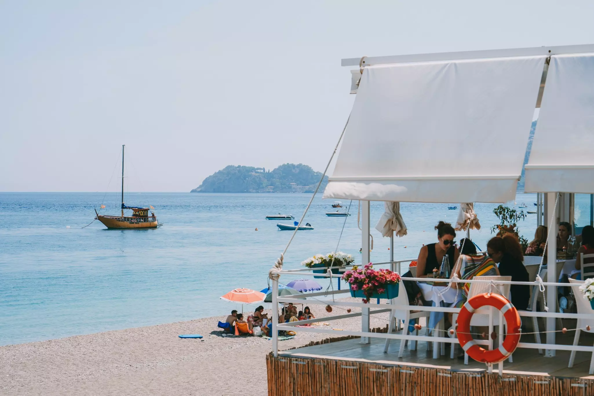 People dine on the terrace of a restaurant overlooking a beach and boats in the water.