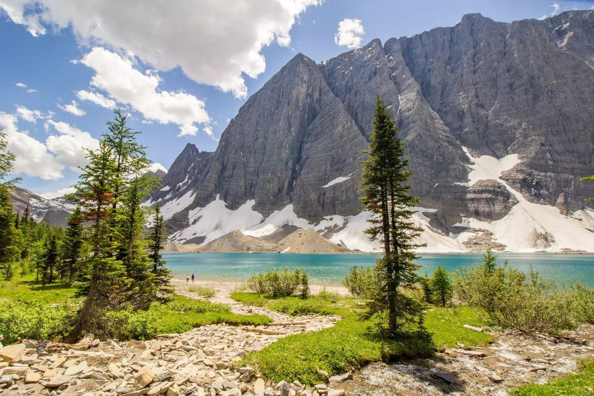 Two hikers stand beside a turquoise alpine lake surrounded by mountain peaks on a sunny day.