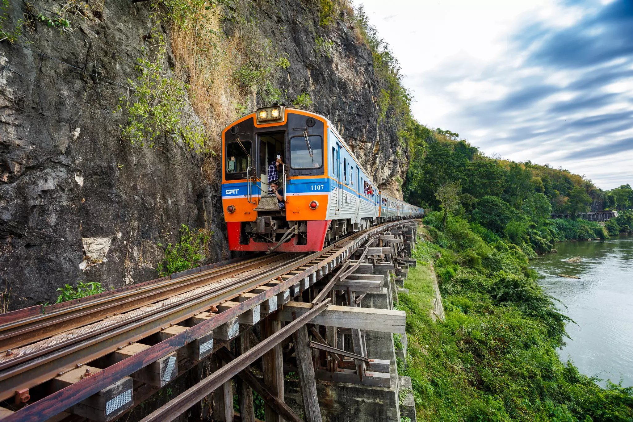 A brightly painted locomotive pulls a train along a trestle next to a riverside cliff.