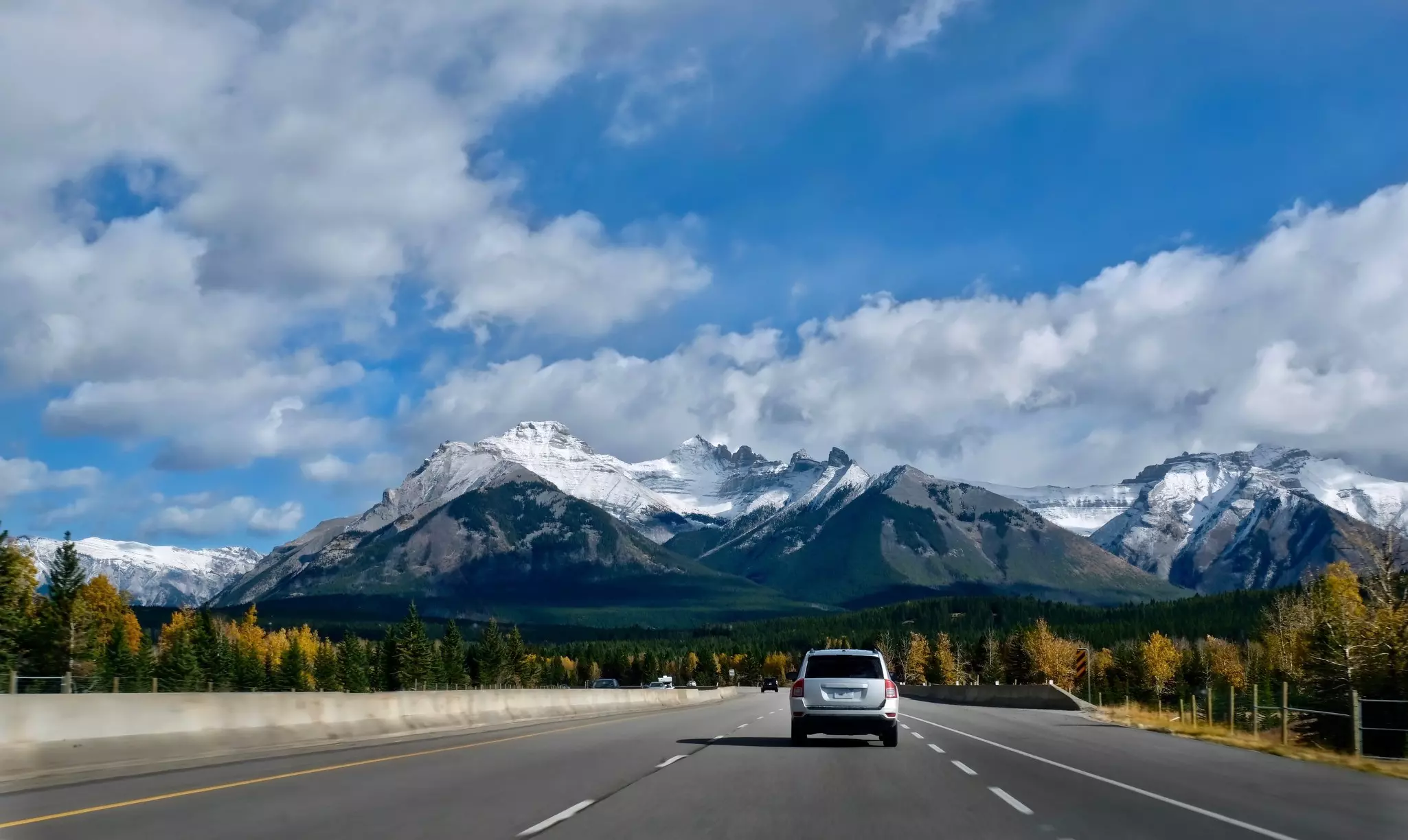 Cars on a highway driving toward snow-covered rocky peaks with clouds and blue sky in the distance on a mostly sunny day.