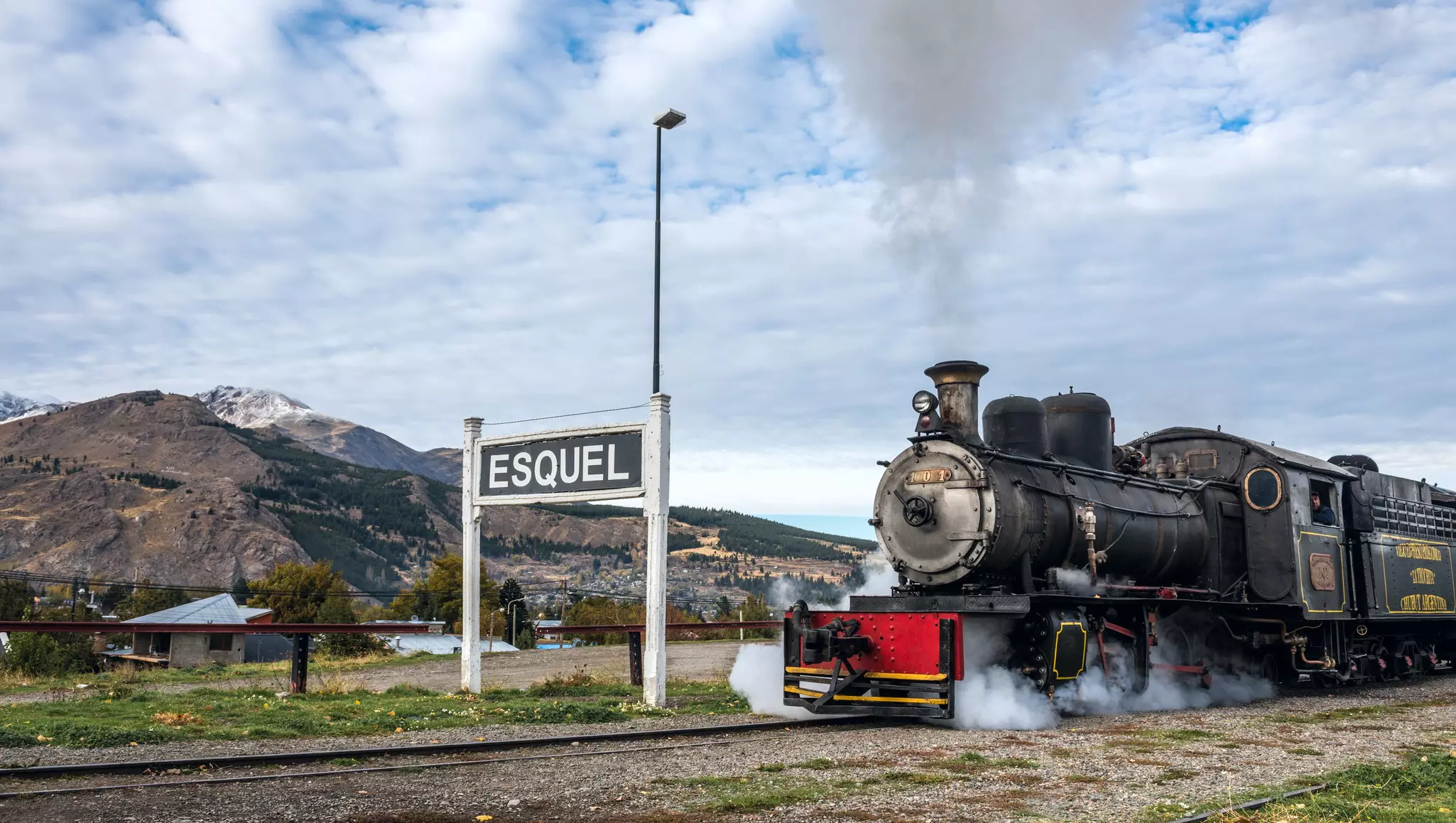 A black steam engine at a train station in a mountainous region near a large sign that reads "Esquel".