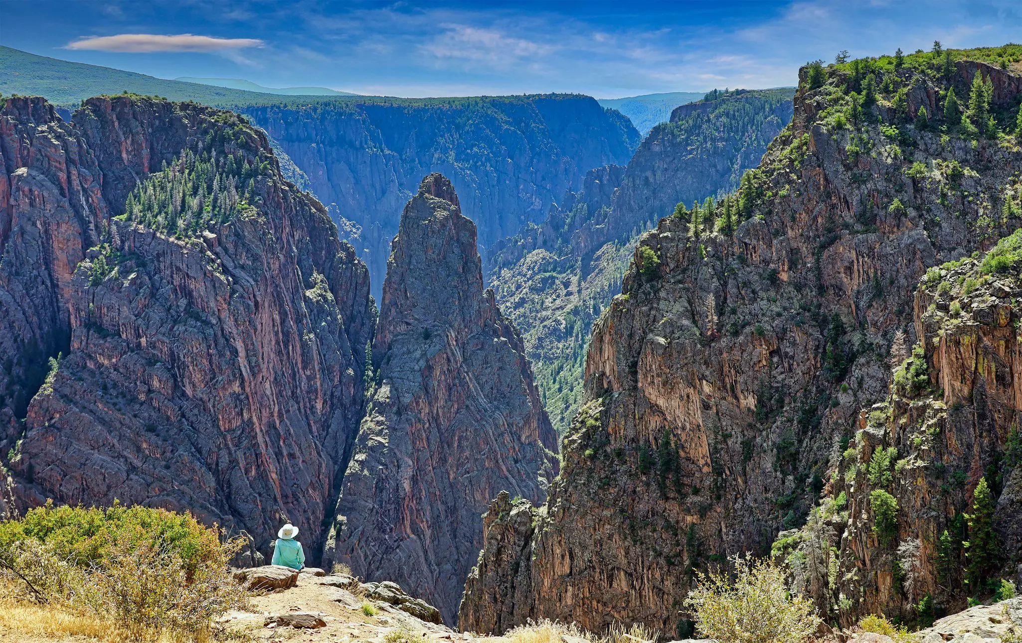 A person sits at a viewpoint looking out over an impressive series of peaks above a canyon.