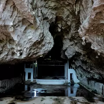 A huge limestone cave overhang with stalactites shelters a whitewashed stage and hall, its wet floor reflecting light