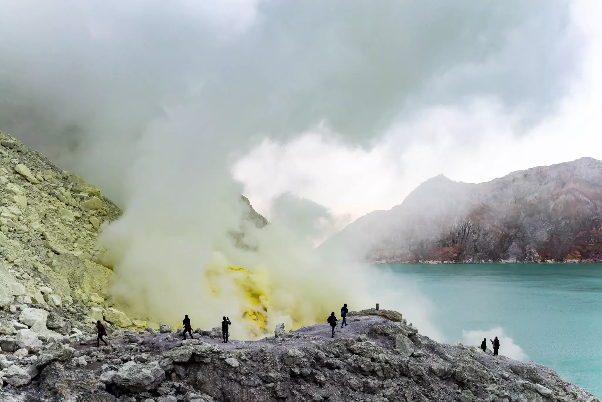 Hikers on a trail through a volcanic landscape approach a crater with blue water and sulfhur clouds rising from the earth 