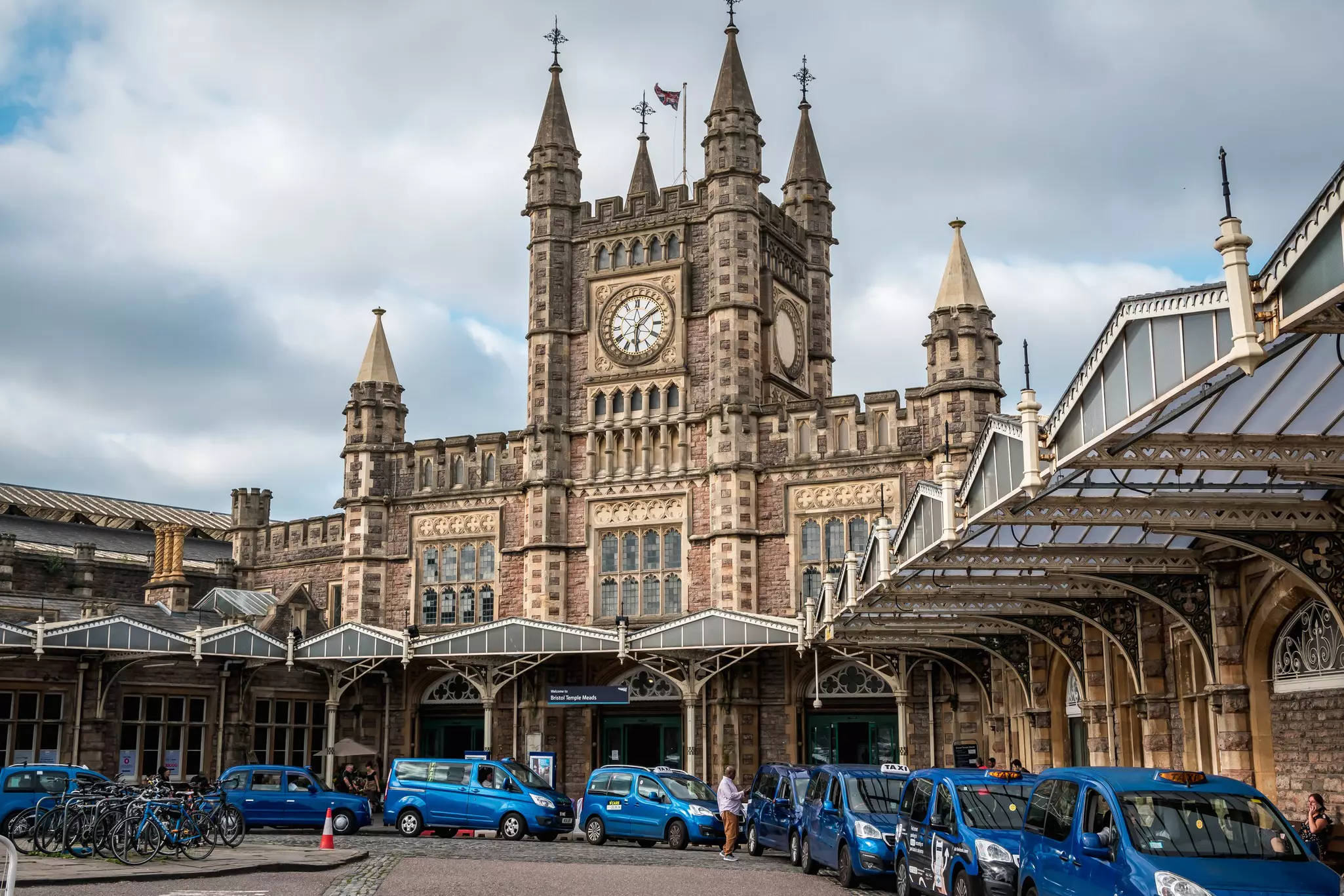 Blue taxis line up outside the grand facade of a train station with a central Gothic clock tower.