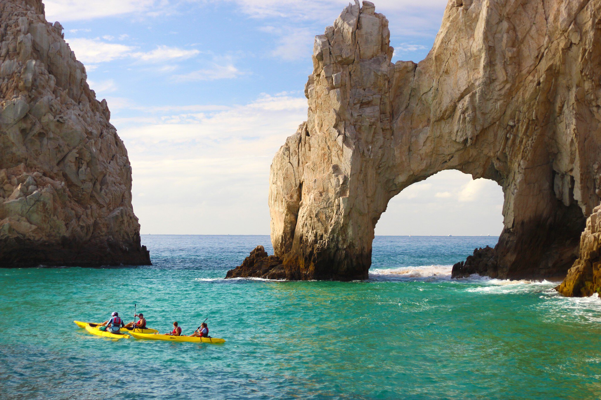 People in yellow kayaks float in turquoise water by a natural stone arch.