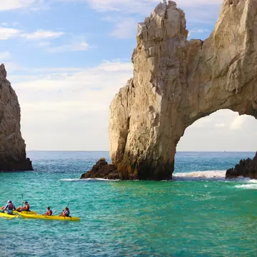 People in yellow kayaks float in turquoise water by a natural stone arch.