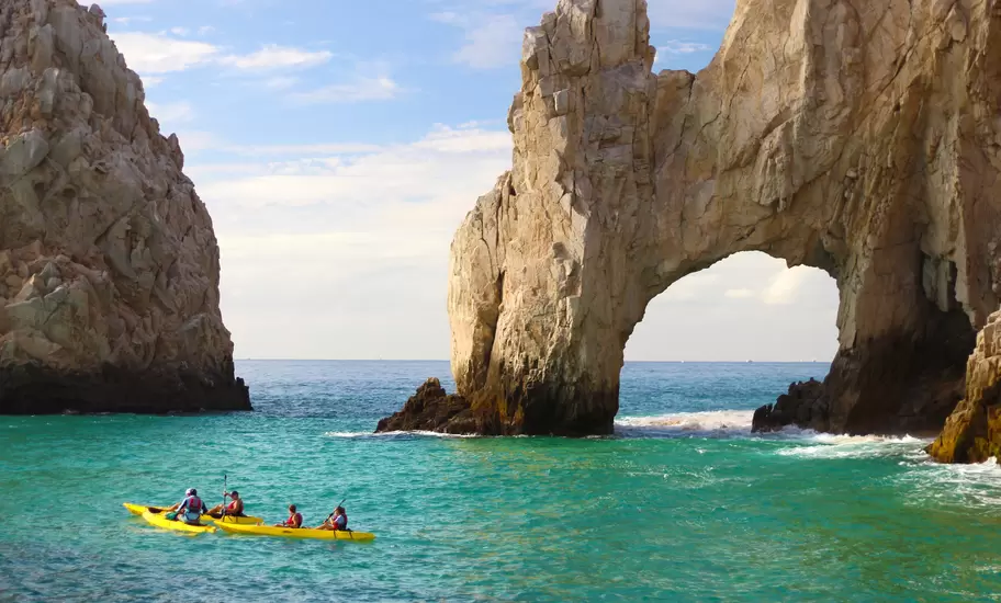 People in yellow kayaks float in turquoise water by a natural stone arch.