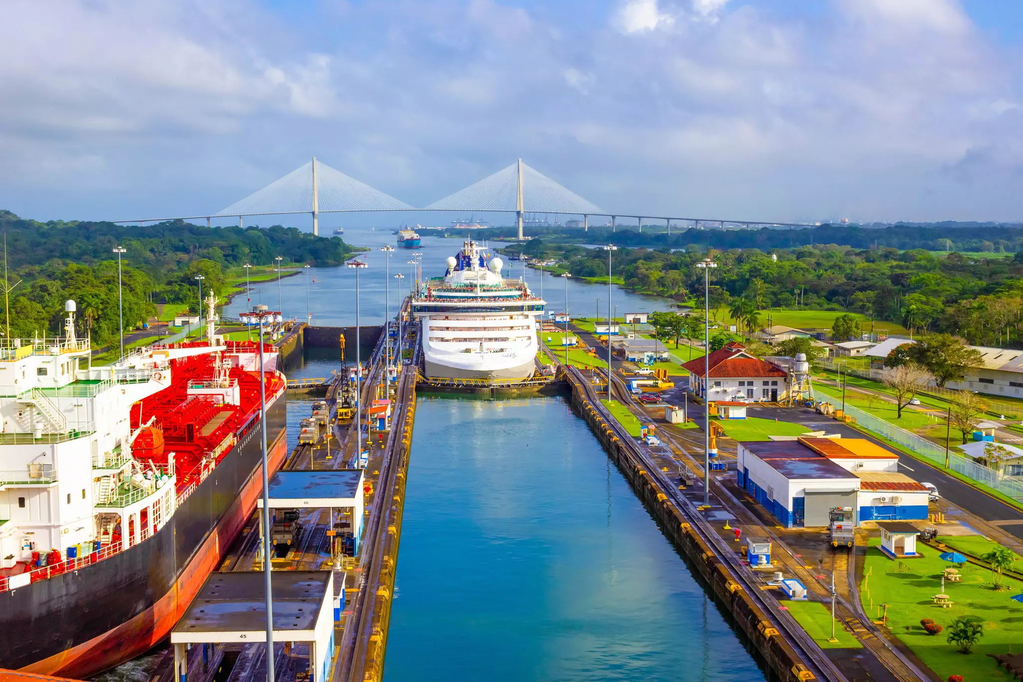 A huge cruise ship is guided through a canal. A large suspension bridge crosses the waterway in the distance.