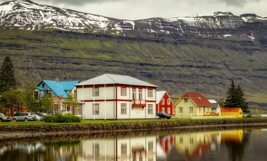 Colourful timber houses reflected in still fjord water below a towering green mountain streaked with snow and gullies, Iceland.