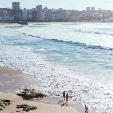 People walking on a sandy beach as white-capped waves roll in, with a row of city apartment blocks along the shore, A Coruña.