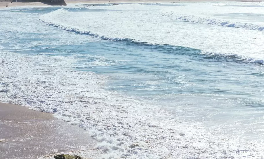 People walking on a sandy beach as white-capped waves roll in, with a row of city apartment blocks along the shore, A Coruña.