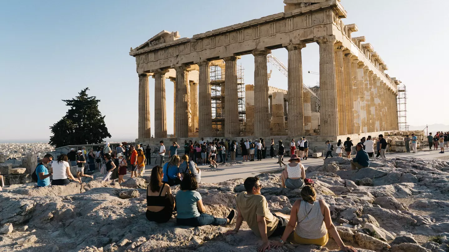 People sit on low rocks or walk along a concrete pathway looking toward a ruin with columns on a sunny day.