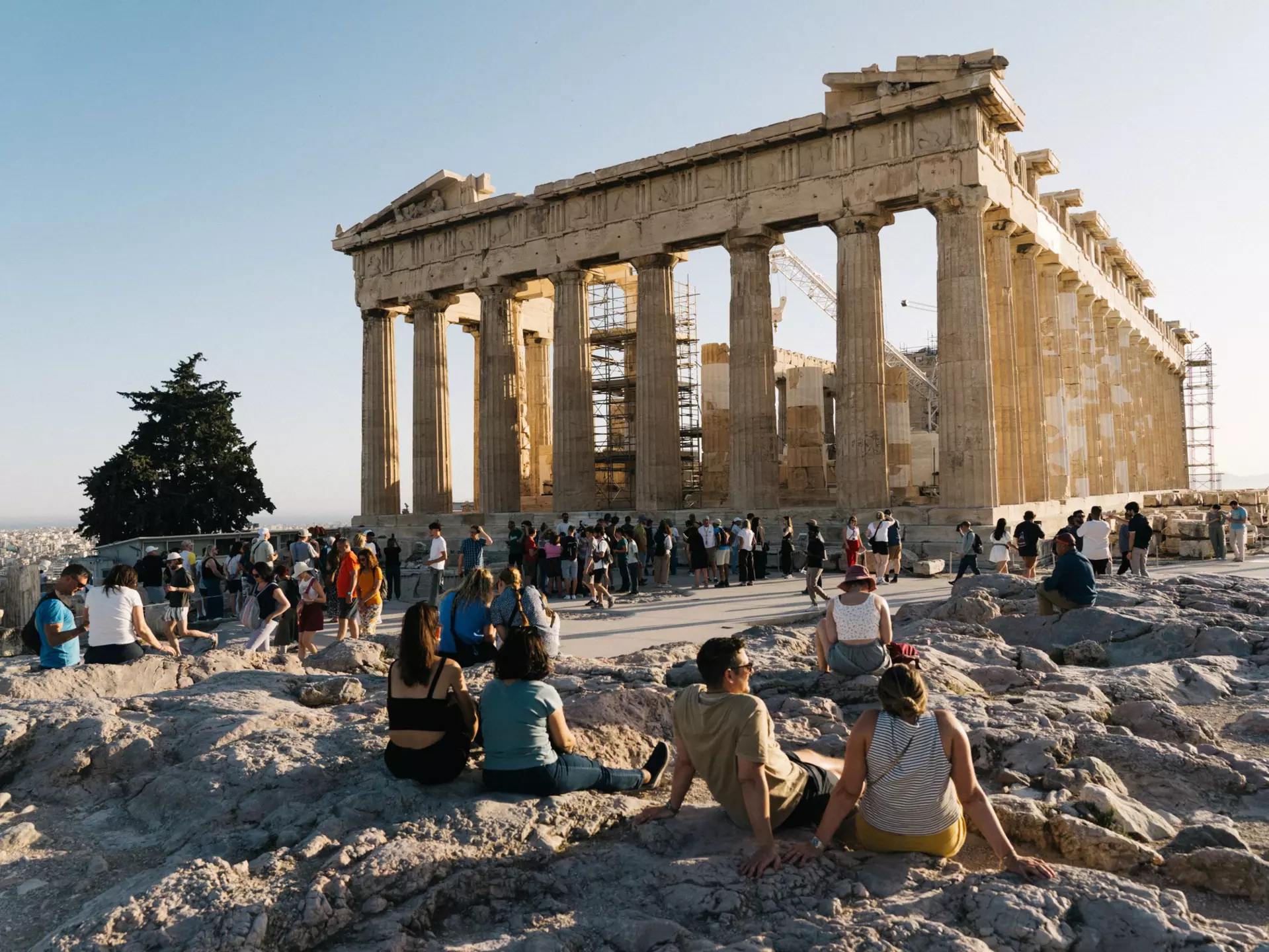 People sit on low rocks or walk along a concrete pathway looking toward a ruin with columns on a sunny day.