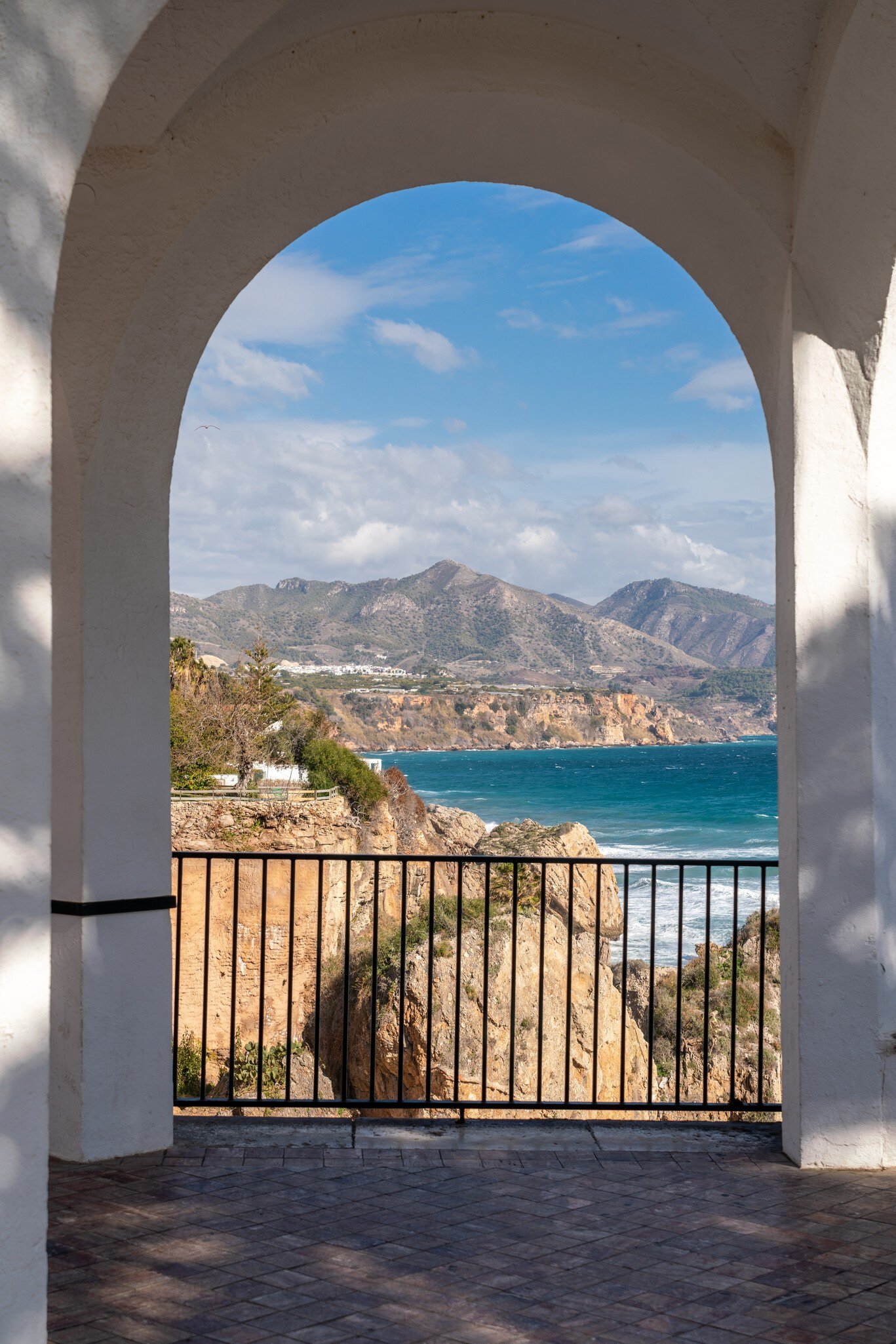 Mediterranean sea, rocky cliffs and mountains seen through a white arch with a railing in Nerja, Spain.