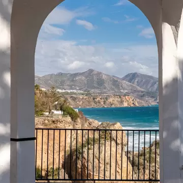 Mediterranean sea, rocky cliffs and mountains seen through a white arch with a railing in Nerja, Spain.