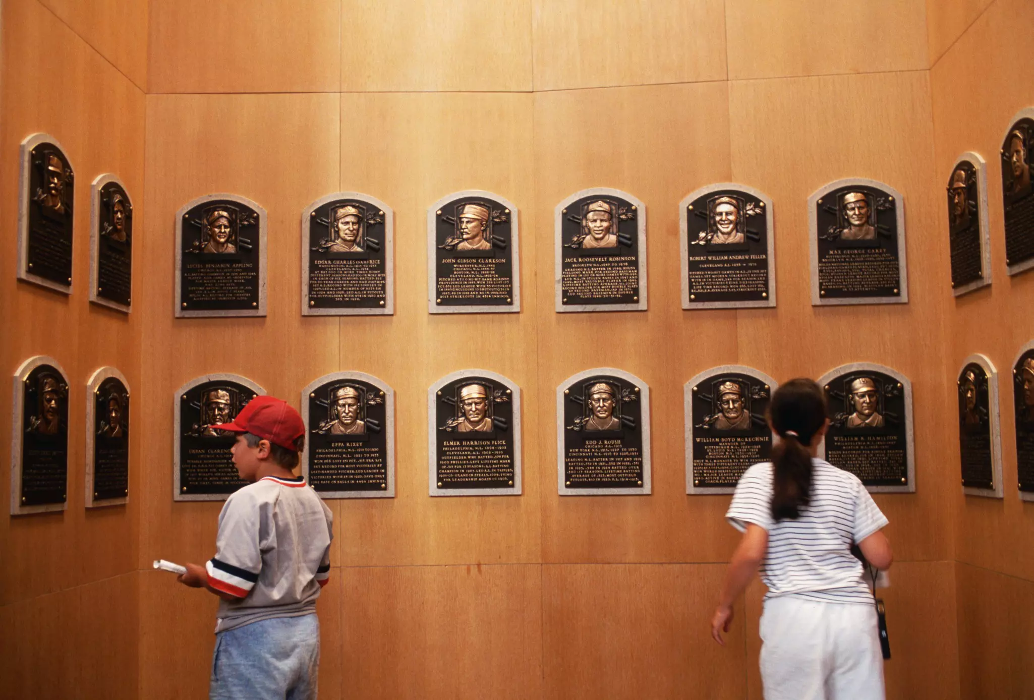 A young boy (who’s wearing a red cap and baseball uniform) and girl look at bronze plaques in a museum.