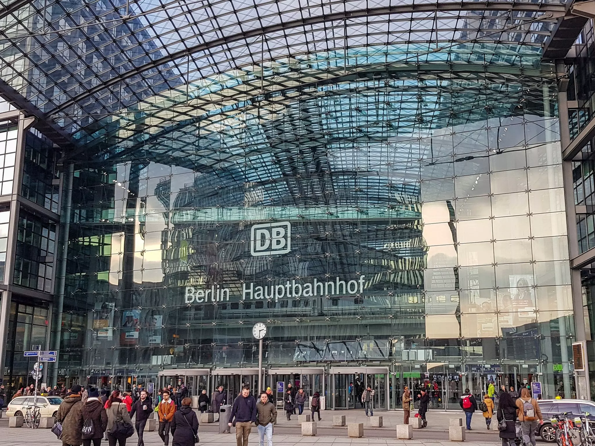 People walking in and out of a modern glass building with the words "Berlin Hauptbahnhof" on the outside.