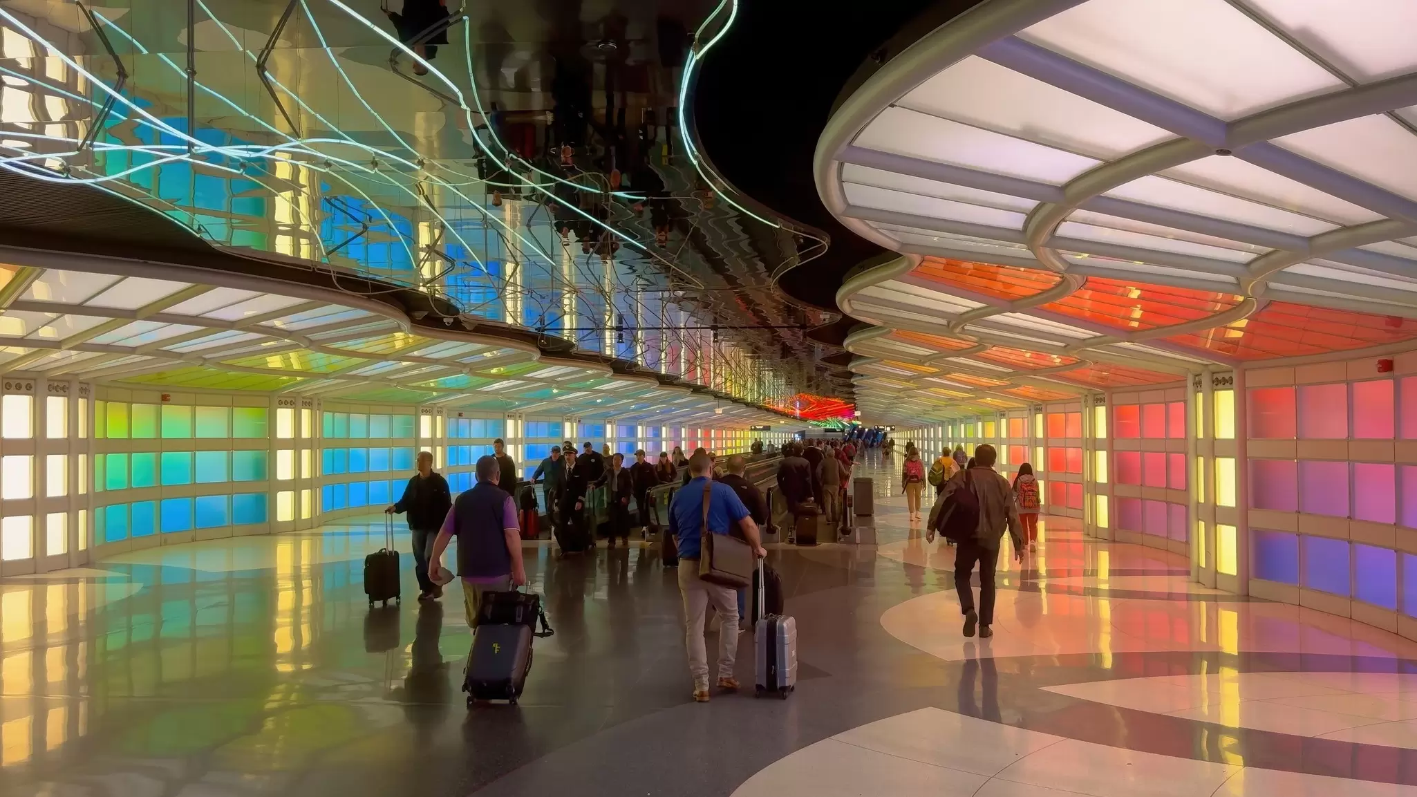 Passengers wheeling luggage walk down a hallway with colorful lighting at Chicago O'Hare Airport's modern terminal 