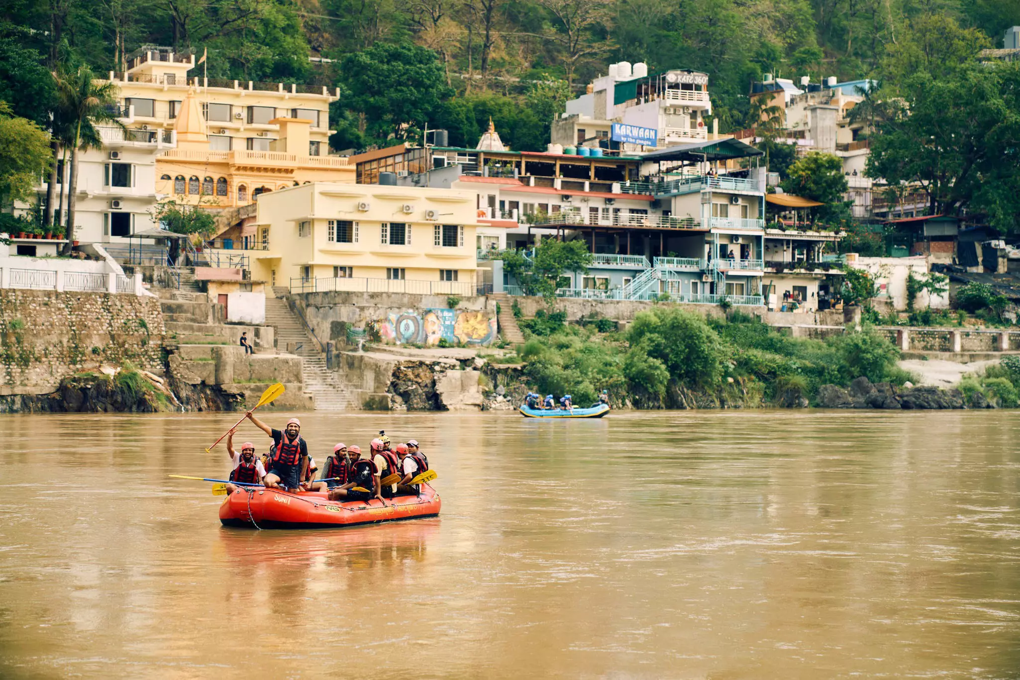 A group of people in a raft on a river with pastel-colored buildings and trees in the background