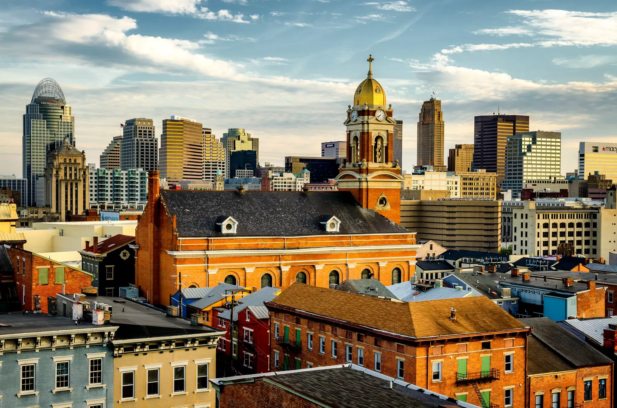 A gold-domed brick church rises above rooftops with the downtown Cincinnati skyline behind at golden hour.