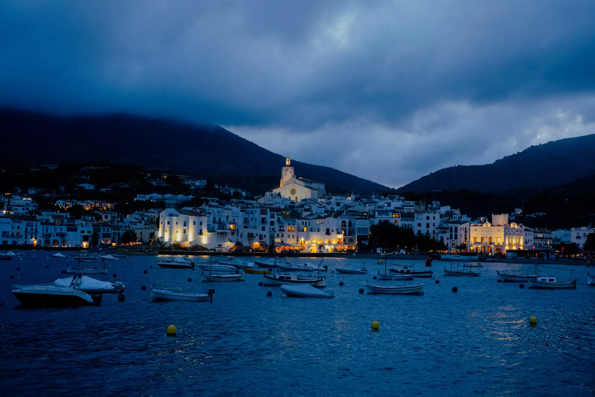 A waterfront town at night with a few lights on and boats in the water. 