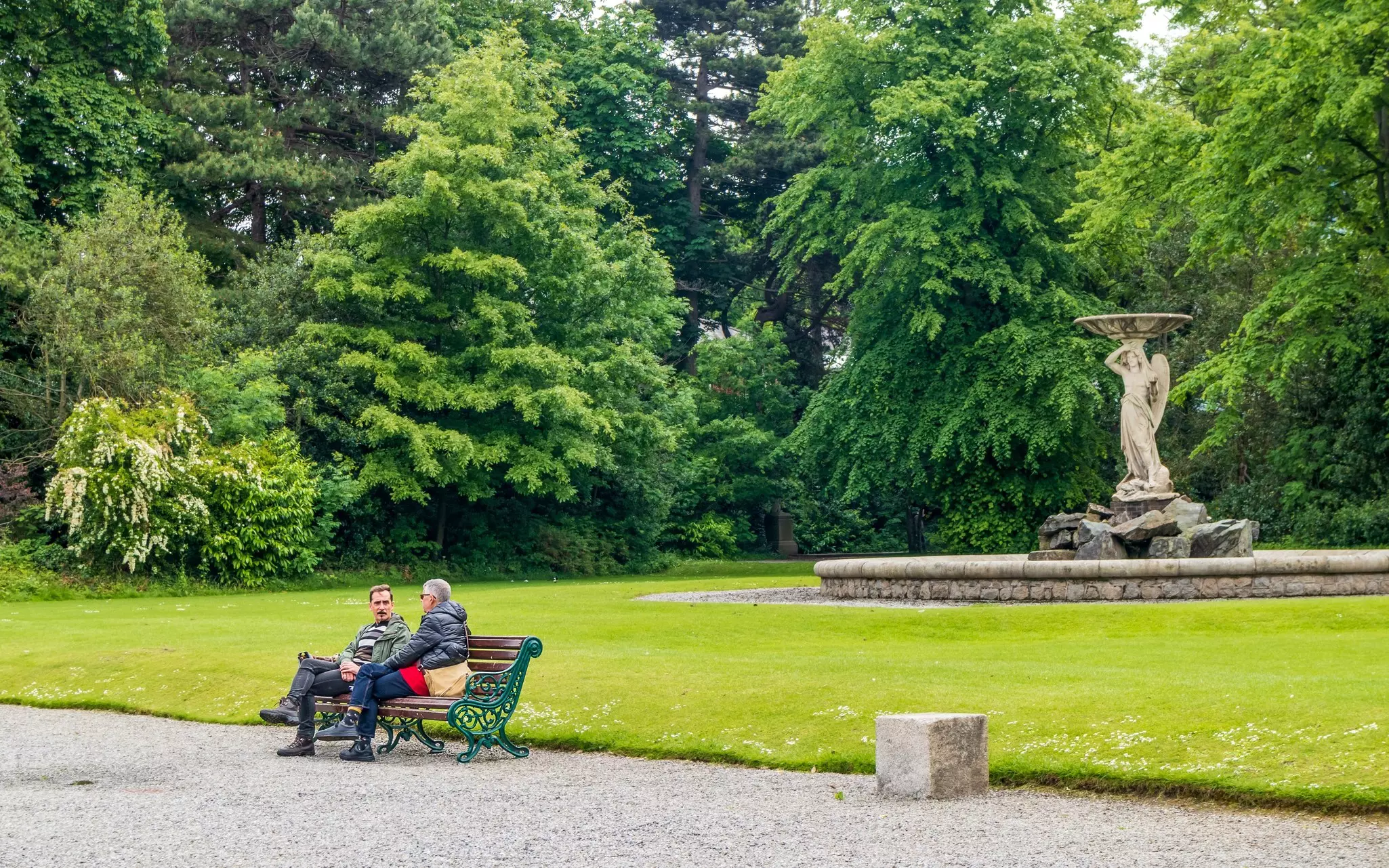 Two people sit on a bench by a lawn in an elegant city parkl.