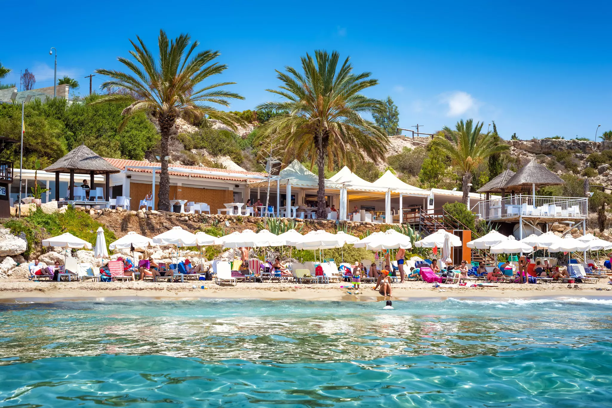 A shot taken from the sea showing people relaxing on sun-loungers and under parasols on a sandy beach backed by a restaurant with a large terrace