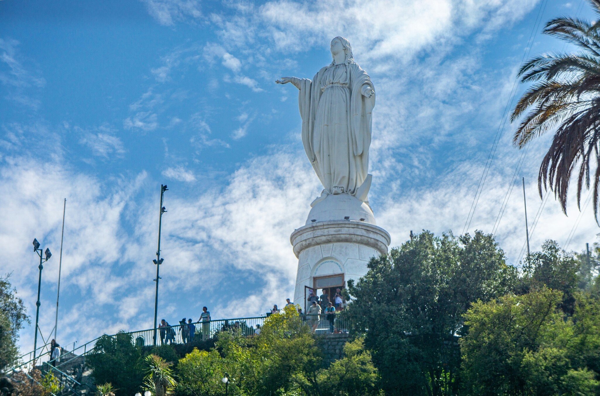 A huge statue of the Virgin Mary at the top of a hill in a city park, with people standing at an overlook in front.