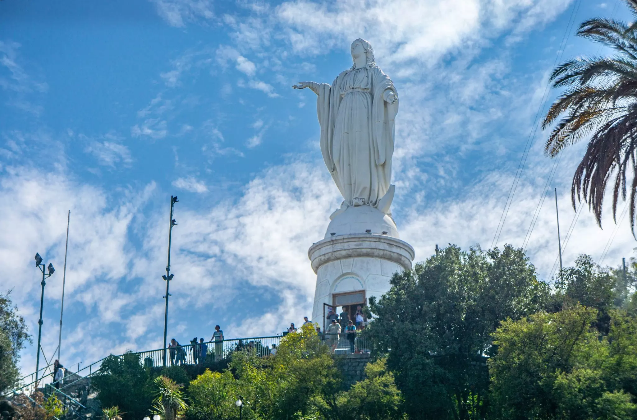 A huge statue of the Virgin Mary at the top of a hill in a city park, with people standing at an overlook in front.